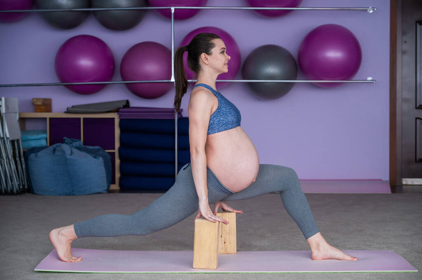 Pregnant flexible European woman is engaged in fitness. The expectant mother in the last trimester of pregnancy practices yoga with the help of yoga blocks. - Foto, immagini
