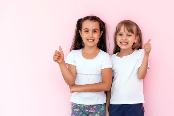 Children showing like. Two adorable happy little girls gesturing thumbs up together and smiling to camera, excellent feedback, good job. indoor studio shot isolated on pink background - Photo, Image