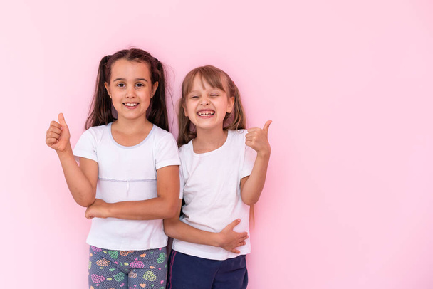 Діти показують, як. Дві чарівні щасливі маленькі дівчата жестикулюють разом і посміхаються до камери, відмінні відгуки, хороша робота. критий студійний знімок ізольований на рожевому фоні
 - Фото, зображення