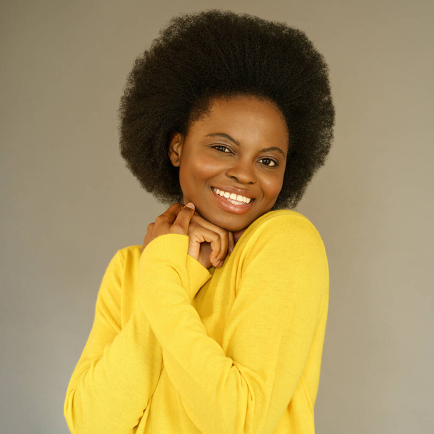 Glückliche afroamerikanische Millennial-Frau mit Afro-Frisur trägt gelben Pullover, der über grauer Wand posiert - Foto, Bild