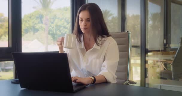Eine Geschäftsfrau in weißer Bluse trinkt einen Drink, während sie im Büro mit großen Fenstern an einem Laptop arbeitet. Tracking Medium Shot, Zeitlupe. - Filmmaterial, Video