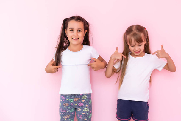 Amazed little kids girls showing white t-shirt, isolated on pink background. Childhood lifestyle concept. Mock up copy space. Pointing index finger aside - Photo, Image