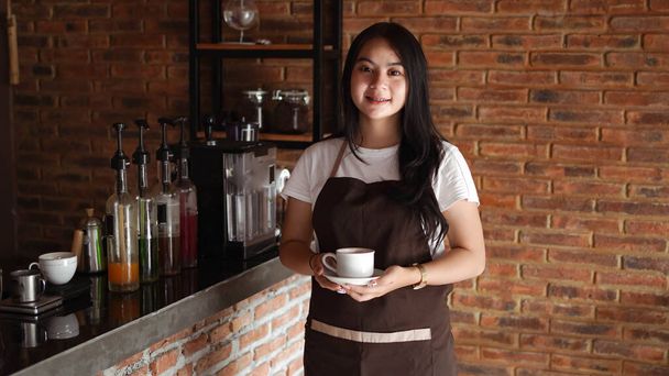 Asyalı Kadın Barista gülümsüyor ve kafe tezgahında kameraya bakıyor. Kahveci kadın kafede kahve bardağı tutuyor. - Fotoğraf, Görsel