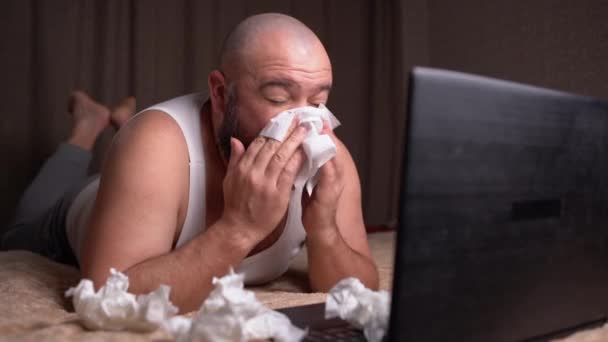 Ein wohlgenährter bärtiger Mann in lässiger Kleidung liegt zu Hause auf der Couch und pustet Rotz in ein Taschentuch. Schnupfen und Allergie-Konzept - Filmmaterial, Video