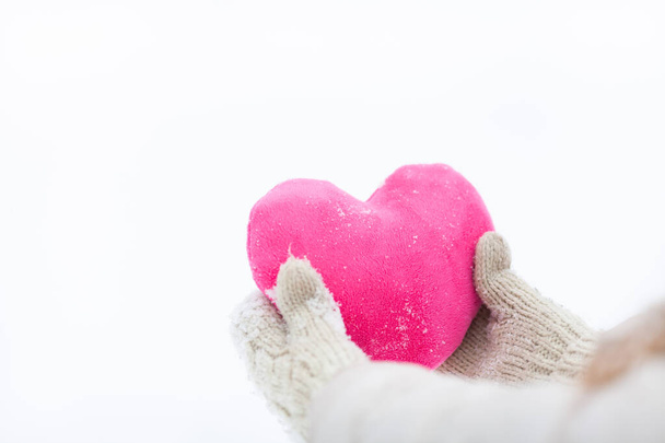 Handen van het meisje in warme gebreide handschoenen met een roze hart op een witte besneeuwde achtergrond. Liefde winter, cardiale zorg of St. Valentijnsdag concept.  - Foto, afbeelding