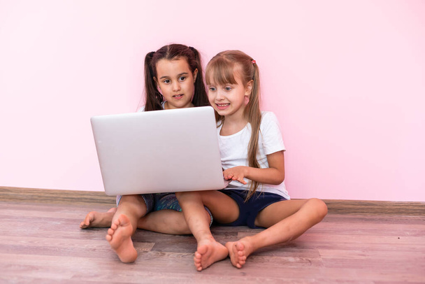 Две девочки работают на ноутбуке, улыбаются и старшая сестра помогает младшей - Фото, изображение