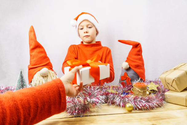 Το παιδί λαμβάνει ένα δώρο. Ένα 7χρονο αγόρι δέχεται ένα δώρο καθισμένο σε ένα τραπέζι περιτριγυρισμένο από Χριστουγεννιάτικα ξωτικά. Χριστούγεννα και Πρωτοχρονιά. - Φωτογραφία, εικόνα
