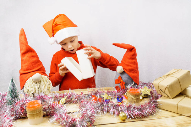 その少年はギフトボックスを見ている。北欧のノームに囲まれた子供は、彼の贈り物を開きます。驚きの感情。クリスマスと新年. - 写真・画像