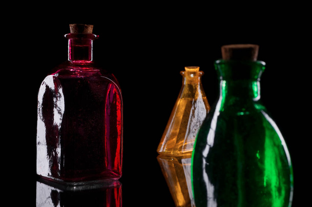 Stillleben dreifarbiger Dekorflaschen mit Korken auf Glas vor schwarzem Hintergrund. - Foto, Bild
