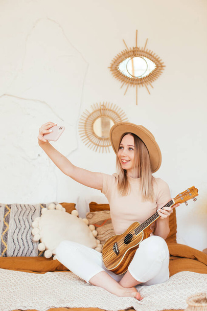 Young funny hipster girl having fun and playing on small ukulele guitar, singing and dancing. wearing a hat, joy, positive mood. instagram live concert. making selfie - Foto, afbeelding