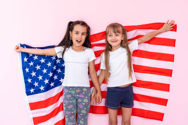 Two adorable little sisters holding american flags on a pink background - Foto, afbeelding