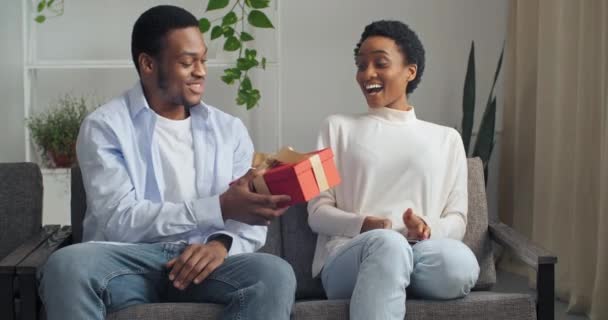 Evdeki kanepede oturan güzel Afro-Amerikan çift. Yakışıklı siyah adam sevgilisine çekici kadın etnik bayan kırmızı hediye paketi veriyor. Doğum gününü kutluyor. - Video, Çekim