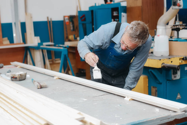 Een oudere meubelmaker in overall en bril schildert een houten bord met borstel op een werkbank in een timmermanswerkplaats.  - Foto, afbeelding