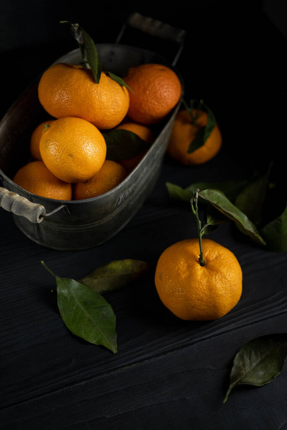 Several tangerines with leaves in a container lie on a black table against a dark background. Rustic style copyspace. - Fotoğraf, Görsel