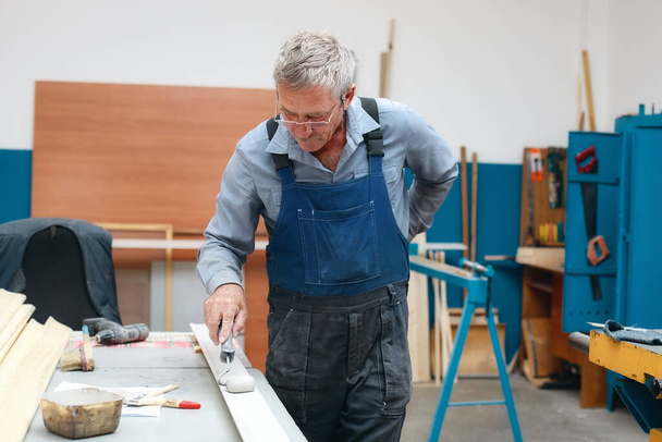 Ein älterer Tischler in Overalls und Brille bemalt eine Holzplatte mit einer Walze auf einer Werkbank in einer Tischlerei. - Foto, Bild