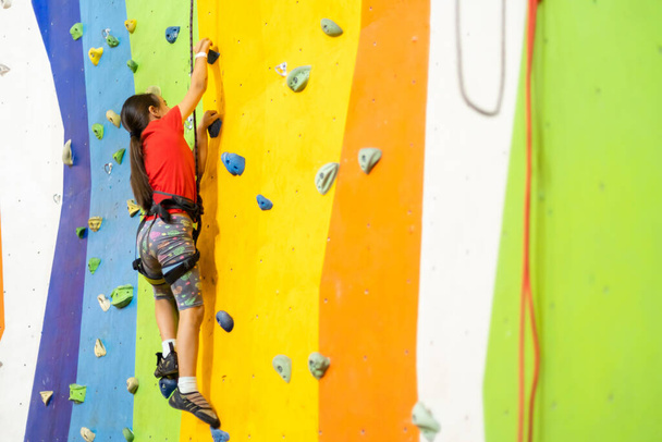Sporty little girl climbing artificial boulder on practical wall in gym - 写真・画像