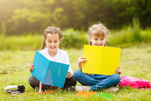 two cute multicultural schoolgirls sitting on lawn under tree and reading book together - Foto, Imagen