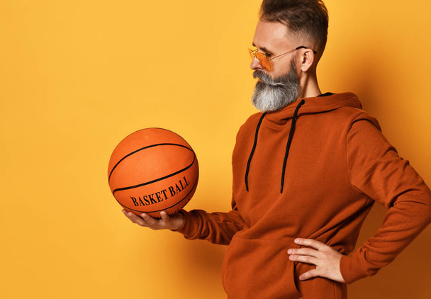 Serious bearded man in sunglasses, hoodie holding basket ball - Photo, image