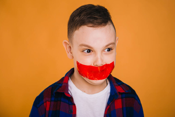 A nine-year-old boy with a mouth taped with red tape. The child is not allowed to speak. Guy rips off the tape from his lips - Foto, Imagen