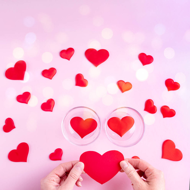 Hands hold a red heart shape, two glasses with hearts and white and red heart shapes on a pink background - Foto, immagini