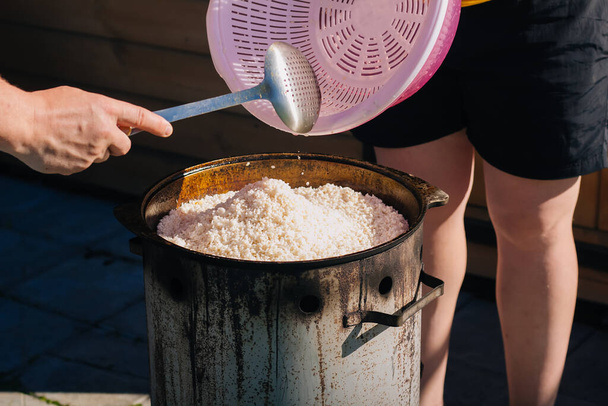το ωμό πλυμένο άσπρο ρύζι χύνεται σε ένα καζάνι. Διαδικασία μαγειρέματος κυμαινόμενο ραφιναρισμένο σιτάρι - Φωτογραφία, εικόνα