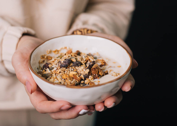 The girl's hands are holding a deep dish with breakfast. Oatmeal, nuts and raisins with milk in a cup. Diet food. Slimming muesli - 写真・画像