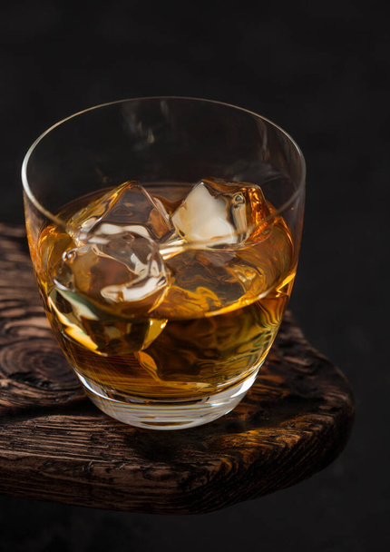 Classic glass of scotch malt whiskey with ice cubaes on top of wooden board and black background - Fotografie, Obrázek