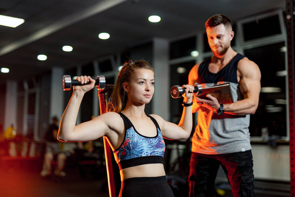 Gorgeous blonde woman trains on sport equipment at a gym. Personal trainer looks at results. - Fotoğraf, Görsel