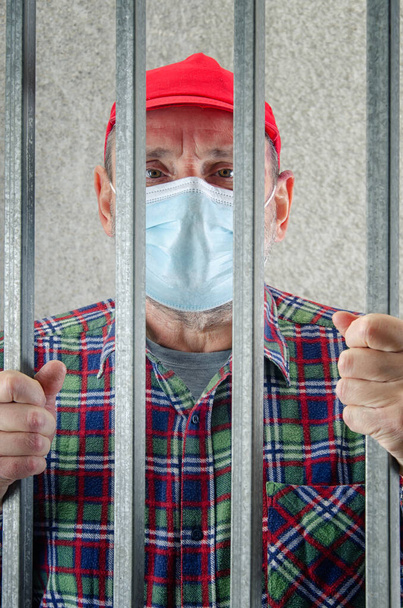 A portrait of an old rebel behind bars. The elderly man is wearing a plaid shirt and a red cap. - Photo, Image