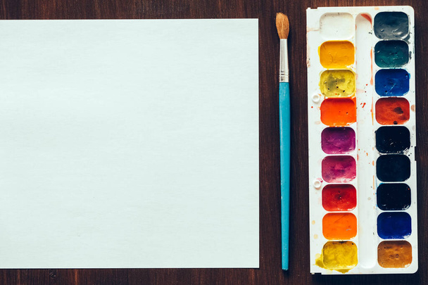 Set Aquarellfarben, Pinsel zum Malen und leeres weißes Papierblatt auf Holzgrund. Ansicht von oben. Kopieren, leerer Raum für Text. - Foto, Bild