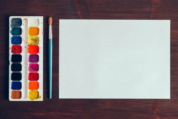 Set Aquarellfarben, Pinsel zum Malen und leeres weißes Papierblatt auf Holzgrund. Ansicht von oben. Kopieren, leerer Raum für Text. - Foto, Bild