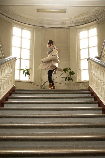 девушка в пальто и сапогах танцует, кружась в зале с окнами и лестницами - Фото, изображение