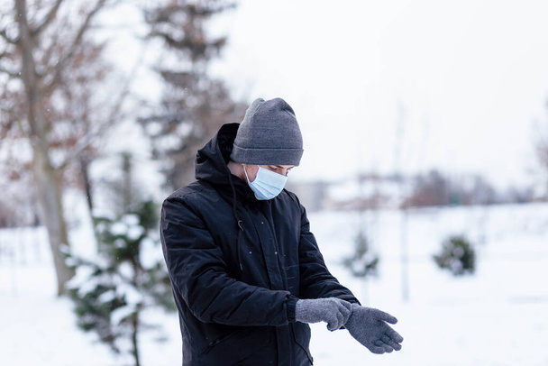 Молодой парень зимой носит маску. Молодой человек в защитной противовирусной маске на улице. Парень в зимней одежде в защитной маске. Молодой человек в теплой одежде и шарфе в зимний день. - Фото, изображение