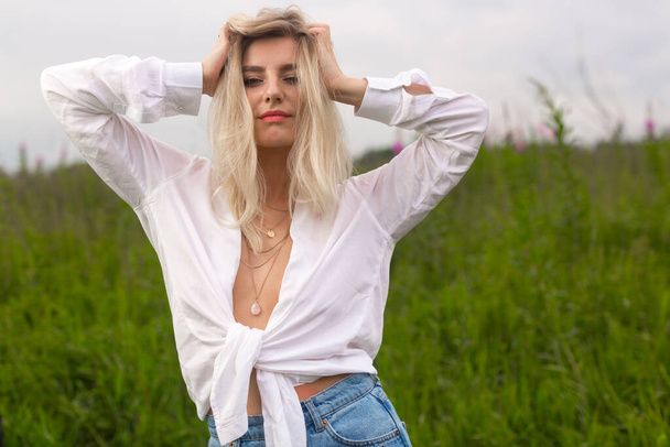 blonde in white shirt straightens her hair against the background of a field - Фото, зображення