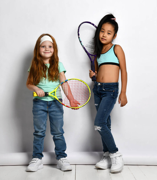 Little tennis player isolated studio portrait shot on white background. Caucasian and Asian girls children in trendy sportive outfit with racket posing for camera. Hobby sport and game activity - Foto, immagini