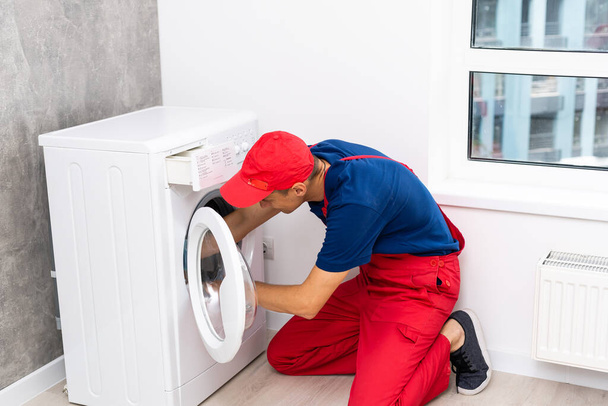 Водопроводчик в комбинезоне с инструментами ремонтирует стиральную машину в доме - Фото, изображение