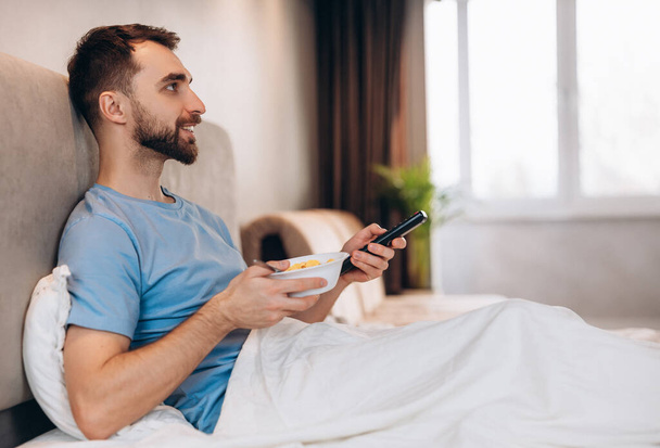 Przystojny młodzieniec z brodą jedzący śniadanie w łóżku i oglądający telewizję. weekend leniwy dzień wolny. - Zdjęcie, obraz