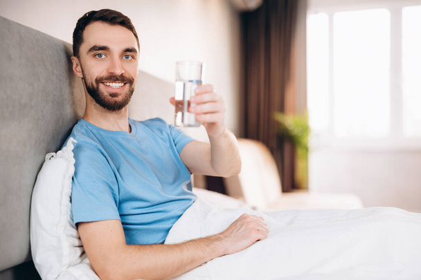 Attraktiver junger Mann mit Bart, der vor dem Frühstück im Bett lächelnd Wasser trinkt, blickt in die Kamera. Faulenzertag am Wochenende. - Foto, Bild