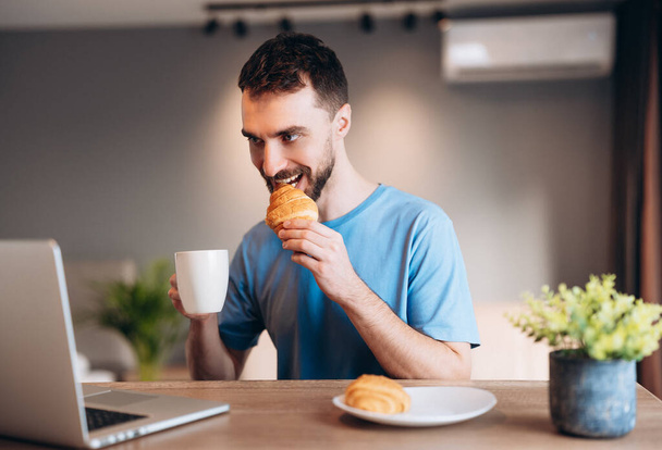 Daily life of young man in kitchen during breakfast using laptop holding a cup of coffee. Attractive male working from home, telecommuting using remote internet job online communication on. - Photo, image