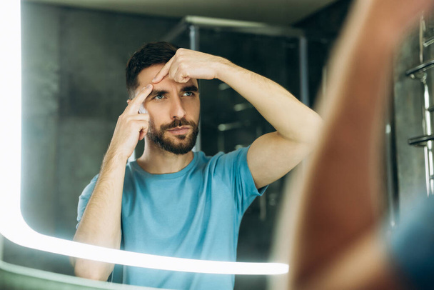 Young man with beard in blue t-shirt squeezing pimple at bathroom mirror acne problem. - Fotoğraf, Görsel