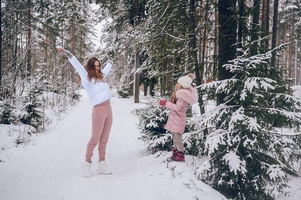 Happy mother and little cute girl in pink warm outwear walking playing snowball fight having fun in snowy white winter coniferous forest with spruce woods outdoors. Family sport vacation activities - 写真・画像