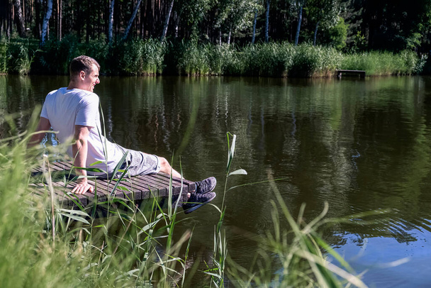 Feliz hombre relajado sentado en el muelle de madera en un lago disfrutando de un día soleado de verano rodeado de vegetación y árboles. Salud mental, reinicio - Foto, Imagen