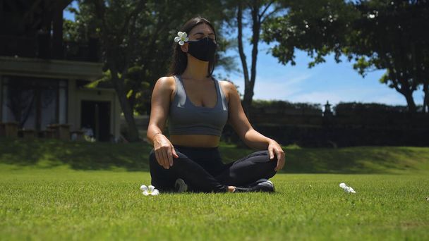 Young European brunette girl practices alone yoga in nature sitting on green grass in lotus pose wearing a black protective mask on her face - Zdjęcie, obraz