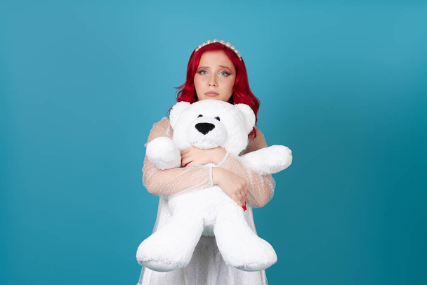 sad, hurt, disappointed, a woman in a white dress with red hair gently hugs a white teddy bear isolated on a blue background - Fotó, kép