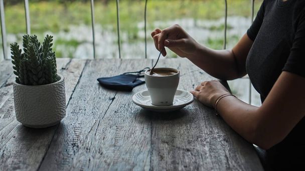 A young European girl sits at a table in a public place with a cup of coffee and nearby to her on a wooden table is a black protective mask - Photo, Image