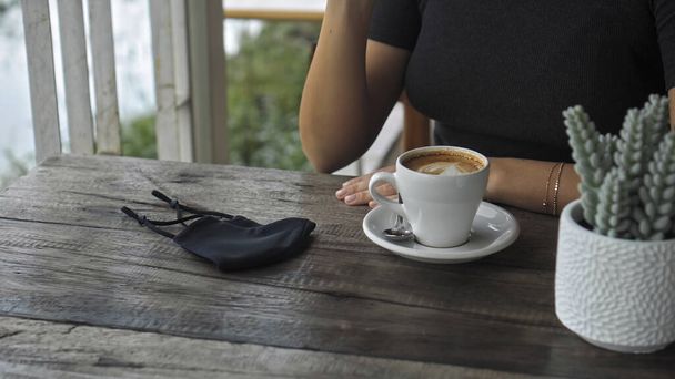 A young European girl sits at a table in a public place with a cup of coffee and nearby to her on a wooden table is a black protective mask - Zdjęcie, obraz