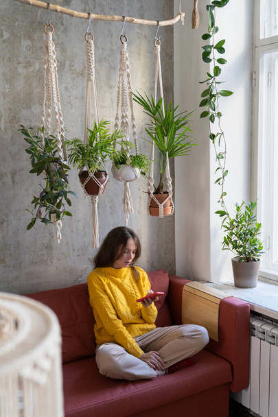 Femme pigiste en pull jaune, au repos, assise sur un canapé sous un cintre en coton macramé avec des plantes d'intérieur, à l'aide d'un téléphone portable. hobby, fait à la main, décoration de maison concept. - Photo, image
