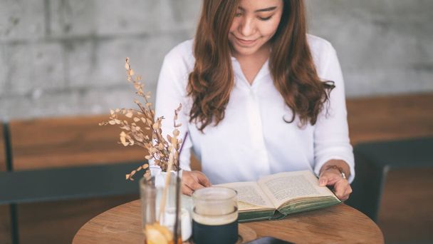 Крупный план красивой азиатской женщины, держащей и читающей книгу, сидящей в кафе - Фото, изображение