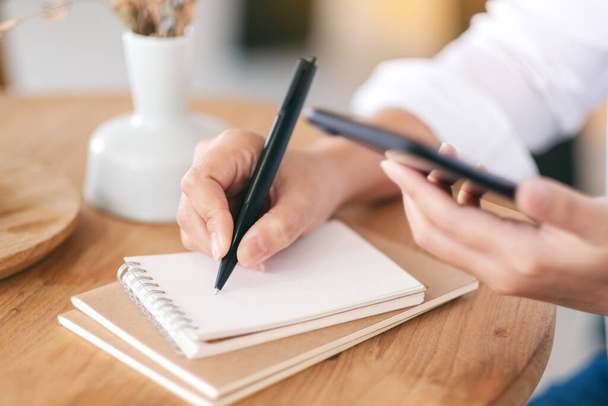 Immagine ravvicinata di una donna che usa il cellulare mentre scrive su un quaderno bianco su un tavolo di legno  - Foto, immagini