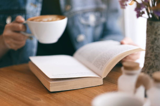 Closeup image of a woman holding and reading a vintage novel book while drinking coffee - Fotó, kép